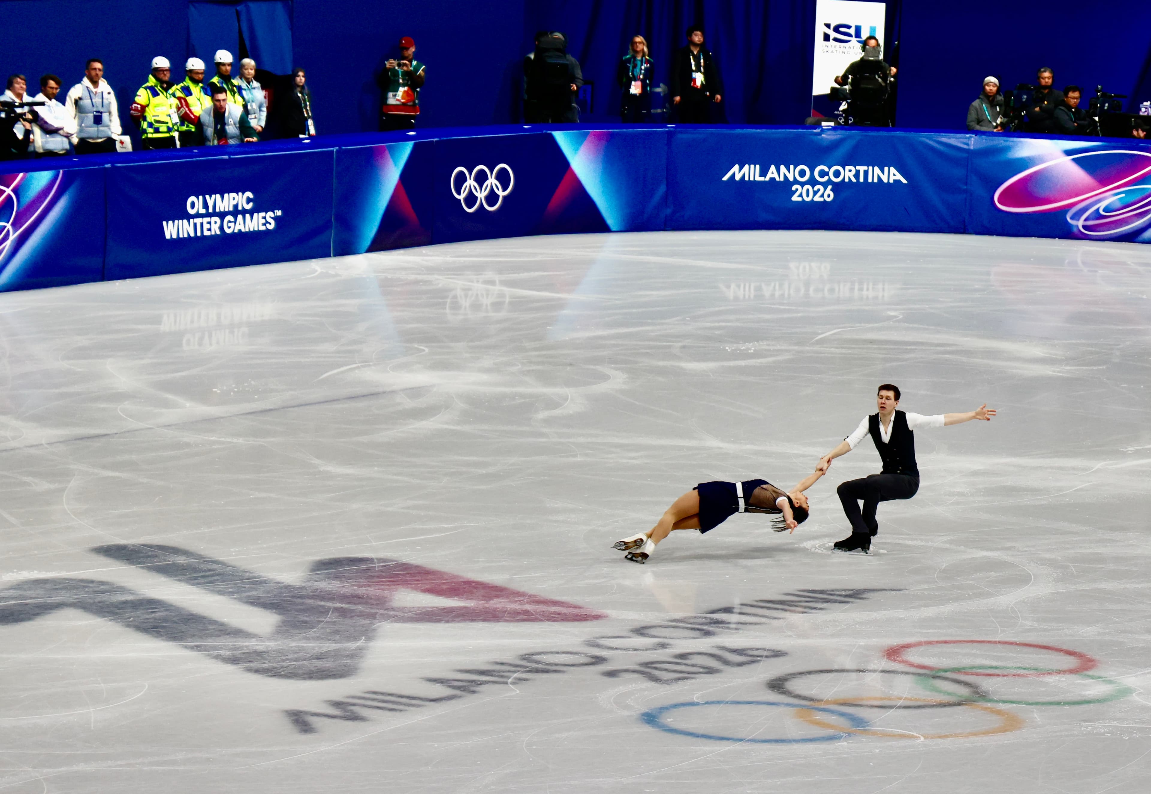 Figure skating pairs team in a dramatic low dip at Milano Cortina 2026, Olympic rings visible