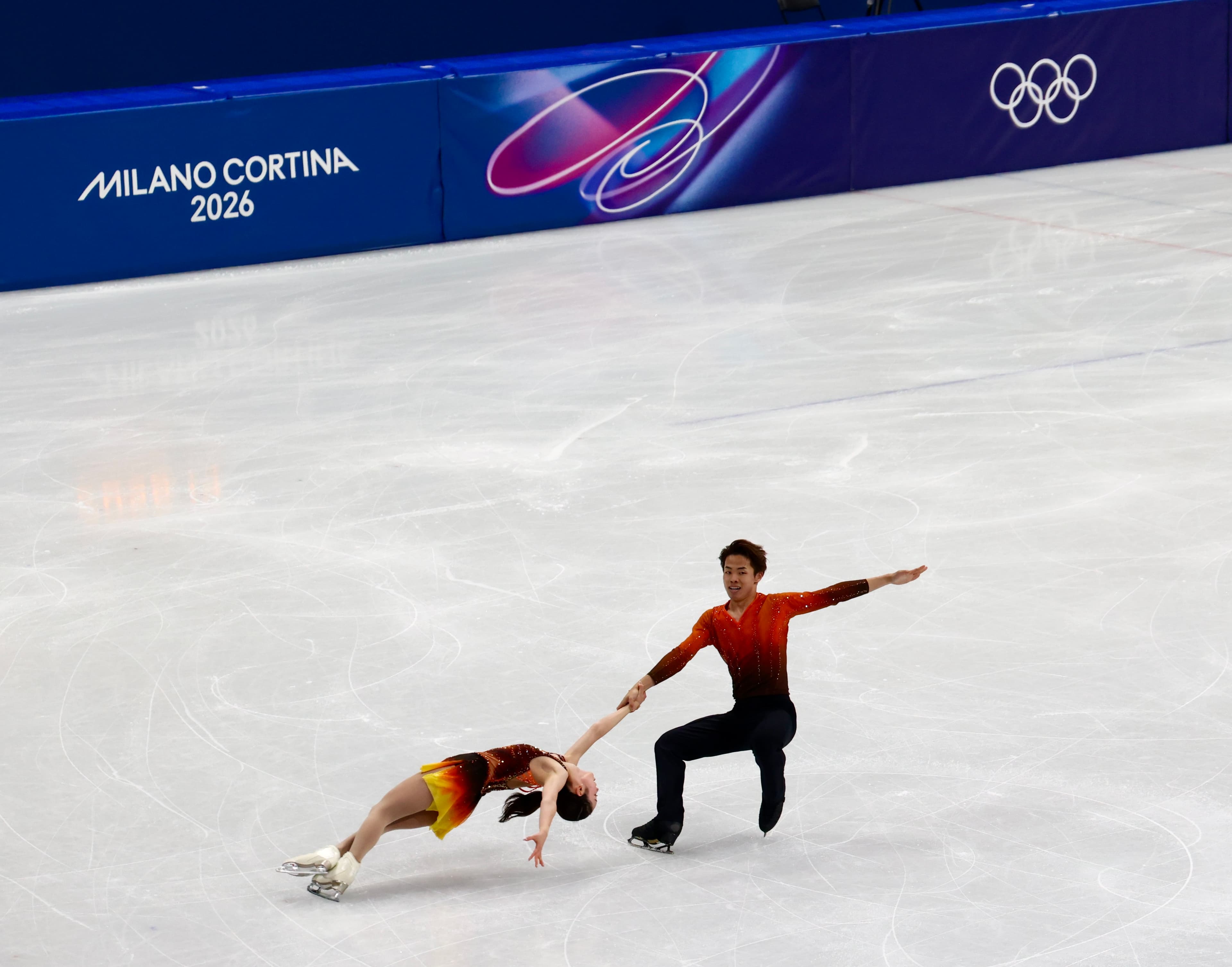 Figure skating pairs in red and orange costumes performing a dip at Milano Cortina 2026