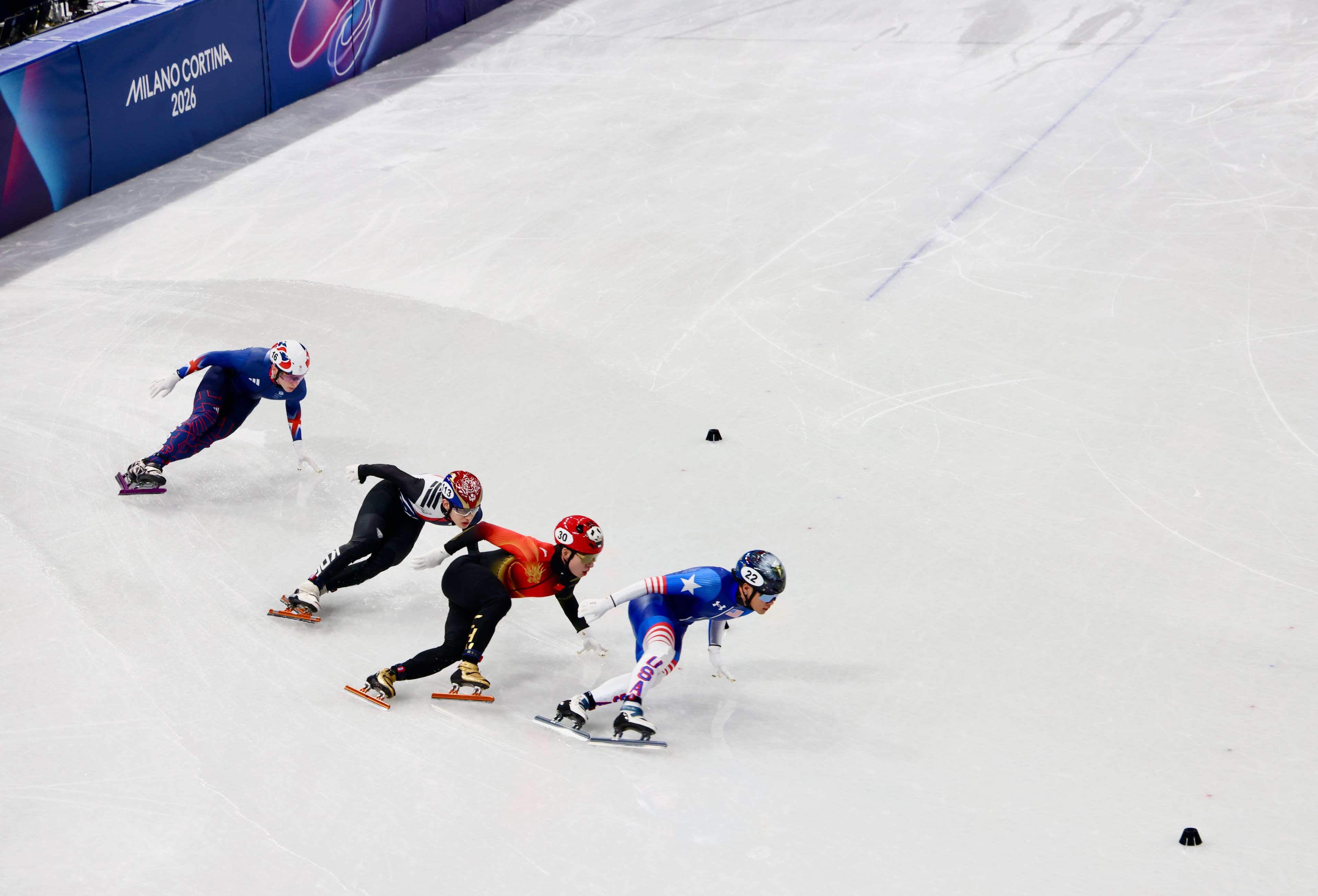 Four short track speed skaters rounding a corner at Milano Cortina 2026