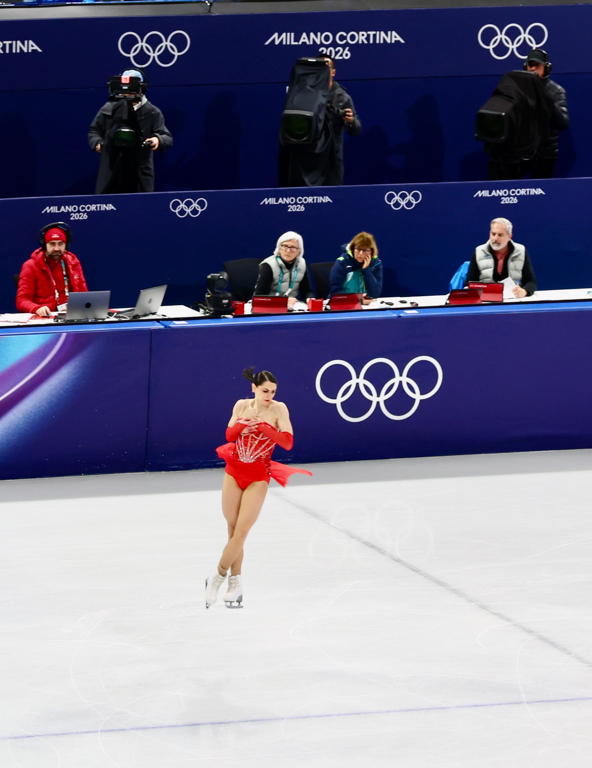 Figure skater in a red dress performing at the Milano Cortina 2026 Olympics with the Olympic rings behind her